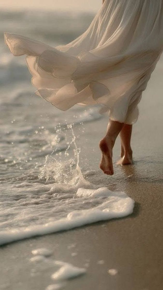 A woman in a flowing white dress walking barefoot through the shoreline